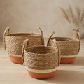 Three woven natural fiber storage baskets with orange-painted bases, arranged on a light wood surface against a neutral beige background.