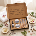 An open kraft gift box containing a small black bottle, two round metal tins, and three slim rectangular boxes, styled on a light marble surface with a folded white towel, dried flowers, crystals, and greenery.