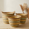 Three woven rope baskets in different sizes arranged on a light beige floor against a cream wall, with dried pampas grass in a vase and part of a wooden chair visible in the background.