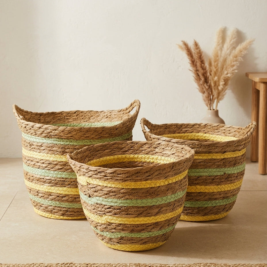Three woven rope baskets in different sizes arranged on a light beige floor against a cream wall, with dried pampas grass in a vase and part of a wooden chair visible in the background.