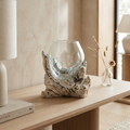 A clear glass bowl vase with a textured, organic beige and gray driftwood base, styled on a light wood console table in a warm, modern interior setting.