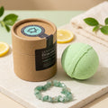 A light green gemstone bath bomb and matching faceted bracelet displayed beside a kraft paper cylindrical package labeled 'Gemstone Braceloo Bath Bomb' on a marble slab with lemon slices and a folded towel in the background.