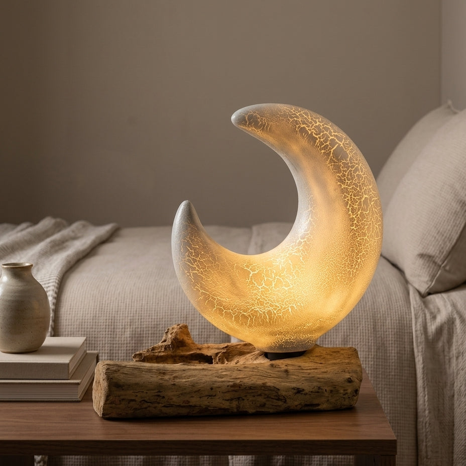 A crescent moon-shaped table lamp with a warm glowing light and crackled glass texture, displayed on a rustic driftwood base on a wooden bedside table in a bedroom setting.