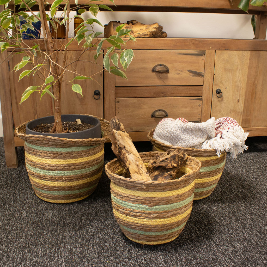 Three woven planters with a plant and towels on a carpeted floor in front of wooden furniture.