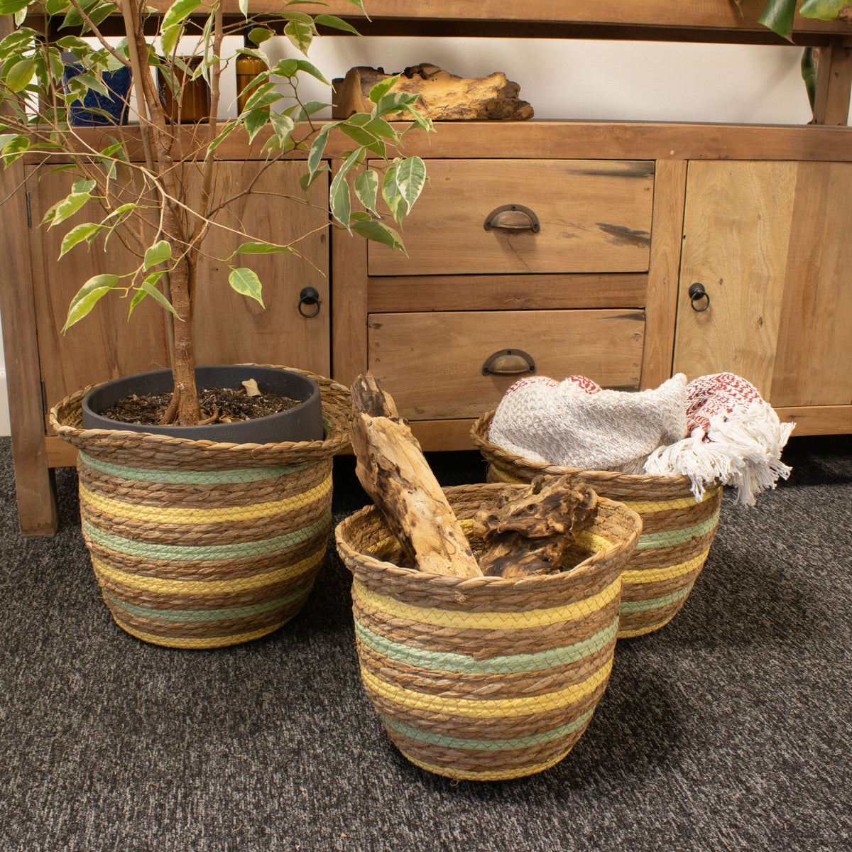Three woven planters with a plant and towels on a carpeted floor in front of wooden furniture.