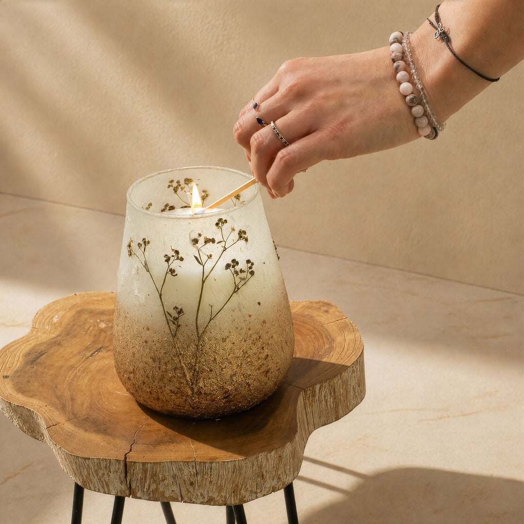 Hand lighting a candle in a decorative glass holder with dried flowers on a wooden surface.